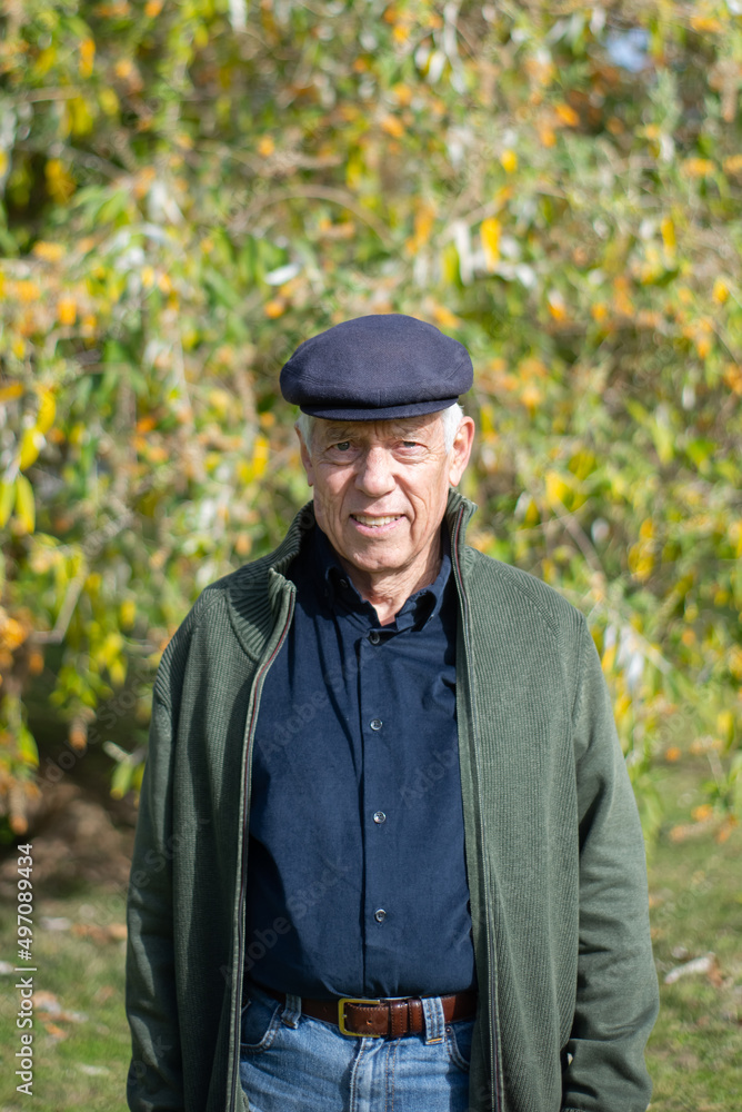 Portrait of elderly man walking in park. Caucasian man in casual clothes and cap resting outdoors standing on green trees background and posing for camera. Active rest of aged people concept