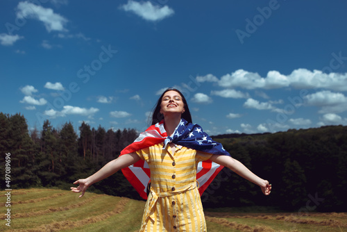 Happy 4th of July! Independence Day celebrating. Patriotic woman hold wrapped in american national flag waving on wind and walking on the field. Stars and stripes. Freedom concept. 