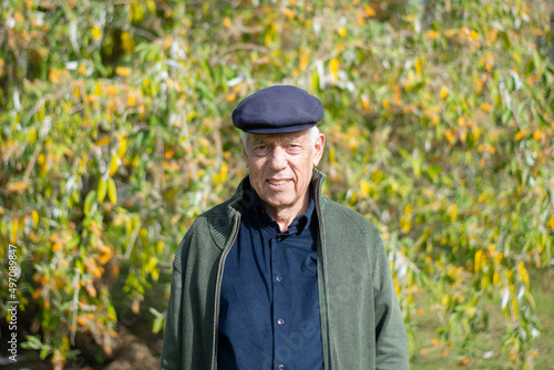 Portrait of aged man resting in spring park. Cheerful man in casual clothes and cap spending time outdoors standing on green trees background looking at camera. Leisure activity of aged people concept