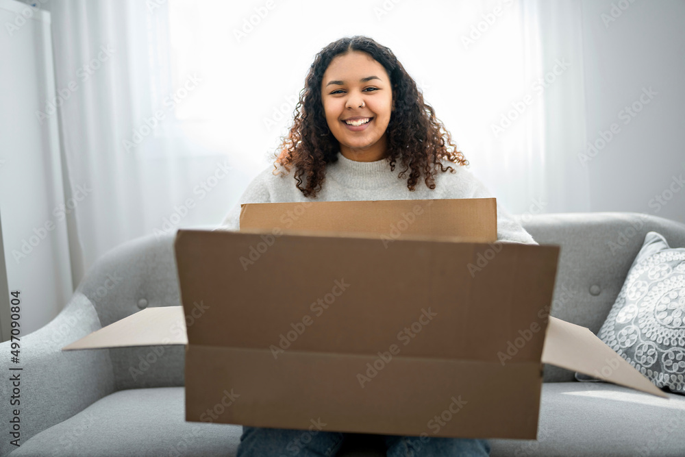Excited young woman unpacking huge carton box sit on sofa at home Excited young woman unpacking huge carton box sit on sofa at home