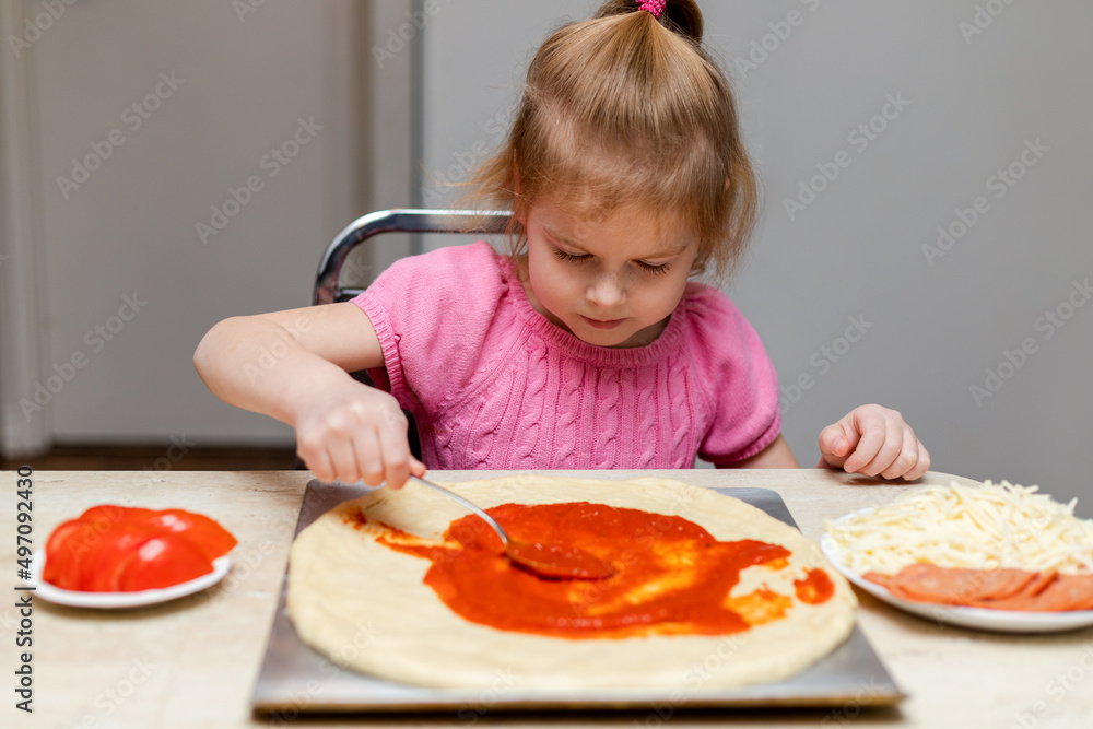 Little child making pizza, sitting at table in kitchen at home, putting pizza sauce on dough. Small girl cooking food.