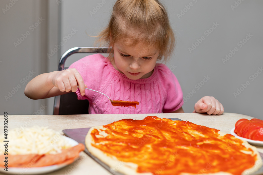 Small child cooking at home sitting at the table. Little girl making pizza in the kitchen. kid preparing food, putting sauce on dough Small child cooking at home sitting at the table. Little girl making pizza in the kitchen. kid preparing food, putting sauce on dough