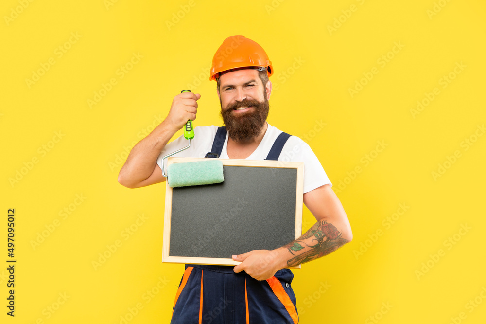 smiling man painter in helmet and work clothes hold paint roller and blackboard with copy space smiling man painter in helmet and work clothes hold paint roller and blackboard with copy space
