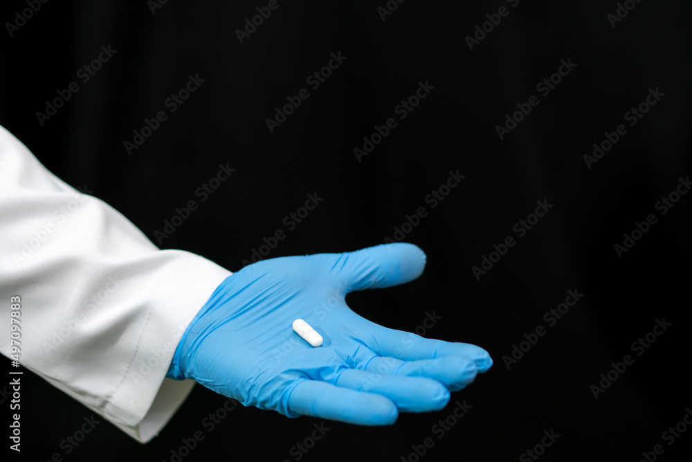A doctor in a white coat holds a pill in his hand on a black background. Medical preparations for the treatment of coronavirus. A young intern doctor is considering a new drug. A doctor in a white coat holds a pill in his hand on a black background. Medical preparations for the treatment of coronavirus. A young intern doctor is considering a new drug.