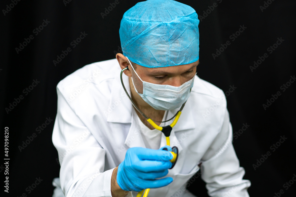 Portrait of a young doctor on a black background. A surgeon in a white coat, a medical mask and a surgical cap holds a stethoscope in his hand. Diagnosis of coronavirus infection and lung disease. Portrait of a young doctor on a black background. A surgeon in a white coat, a medical mask and a surgical cap holds a stethoscope in his hand. Diagnosis of coronavirus infection and lung disease.
