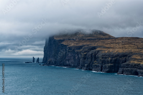 Steep cliff falling vertically into steel-blue ocean with two landmark rocks Risin and Kellingin, top covered with moody cold clouds