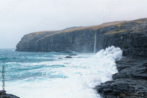 Ice-blue stormy sea breaking to rainy nordic black rocky beach with cliff and waterfall in background covered by misty clouds
