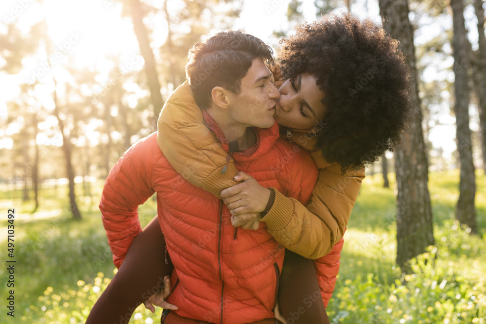 Fototapeta premium Multiracial couple outdoors - Caucasian guy giving a piggyback ride to an African girl in the forest - Concept of tourism, friendship, youth and weekend activities.