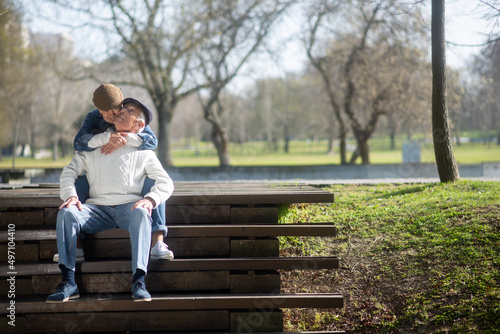Happy elderly couple hugging on stairs in park. Attractive grey-haired woman in cap sitting on wooden stairs behind man hugging his neck and smiling. Love and active rest of aged people concept