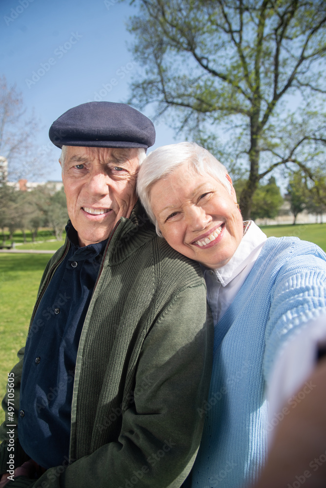 Close-up of elderly couple using phone for selfie. Smiling man and woman standing close woman leaning mans shoulder with her head, both looking at camera. Happiness, active rest of aged people concept