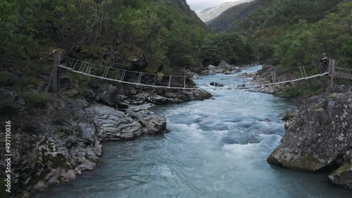 Beautiful scenic drone flight in scandinavia, flowing fast mountain river, big rocks in blue foam water. Footage of adventure inspiring young hiker girl walking on wooden suspension bridge in norway