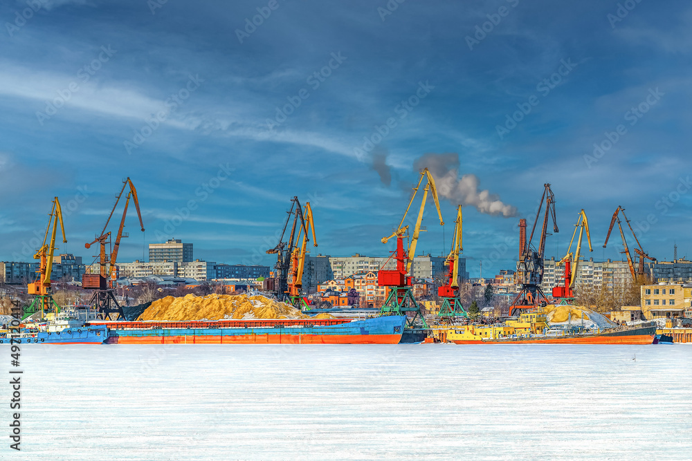 Barges, cranes, ships for the transport of heavy goods, iron ore, sand in the river cargo port in winter. Industrial landscape.