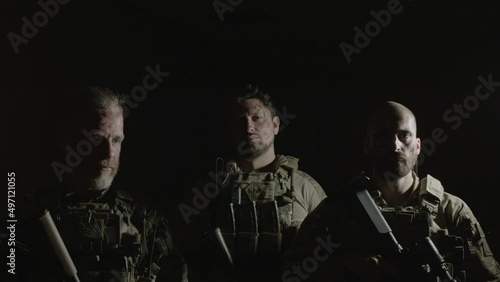 Men in camouflage uniform with guns standing in dark room. Front view of serious friends in bulletproof vests looking at camera, ready to play airsoft in building. Entertainment concept photo