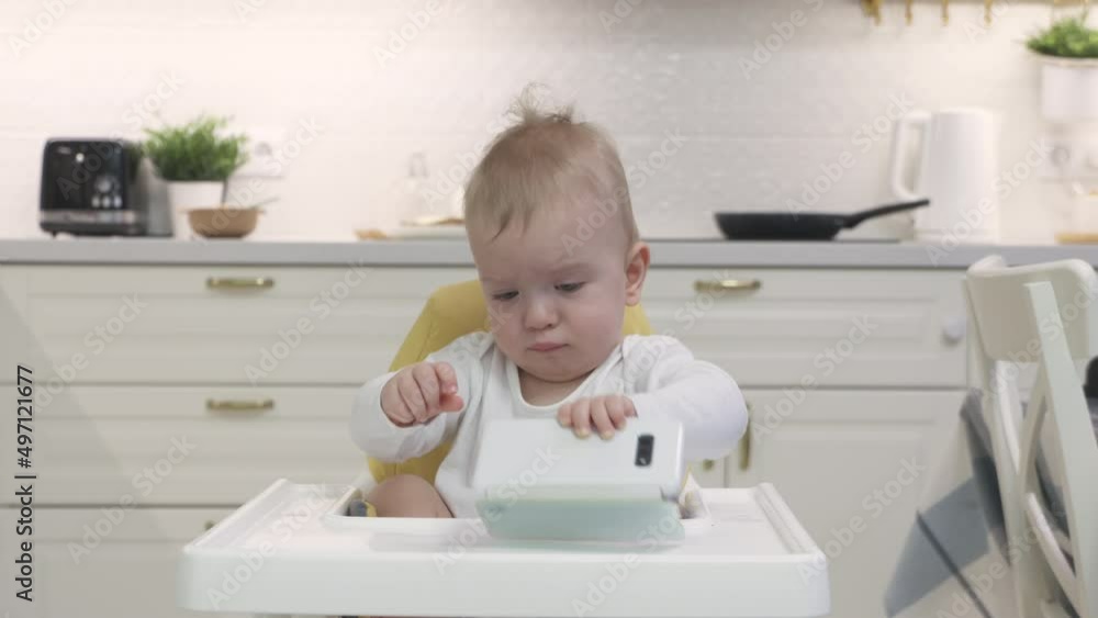 Baby boy sitting on high chair for babies playing with smartphone, cute toddler holding mobile phone, child and modern technology concept. High quality 4k footage Baby boy sitting on high chair for babies playing with smartphone, cute toddler holding mobile phone, child and modern technology concept. High quality 4k footage