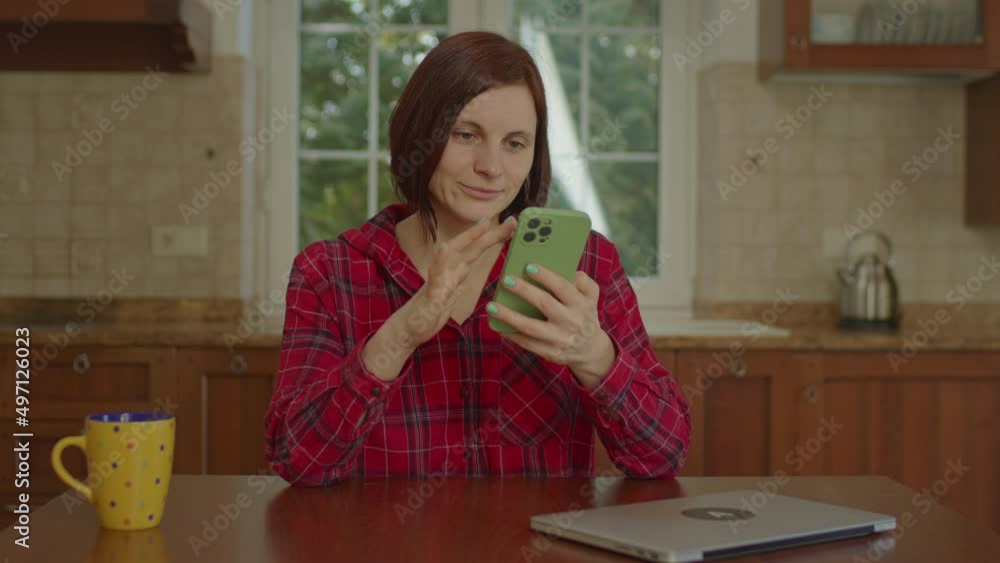 Satisfied 30s woman using mobile phone sitting in kitchen. Female scrolling cell phone screen and smiling. Satisfied 30s woman using mobile phone sitting in kitchen. Female scrolling cell phone screen and smiling.