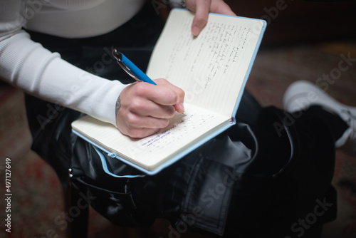 Close-up of young woman taking notes in notebook. Caucasian female student wearing sweatshirt and leather trousers sitting and writing lecture or diary. Education or creativity concept