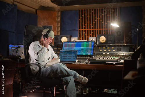 Concentrated young man listening to music sample in headphones. Sound engineer or music producer working at mixing console panel in sound recording studio. Creating music concept