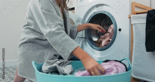 Woman performs household chores in bathroom, laundry room, kneels with wicker basket filled with clothes at washing machine, loads colorful things into drum.