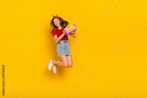 Full body photo of cute teenager girl jump hold present wear t-shirt hairband skirt footwear isolated on yellow background © deagreez