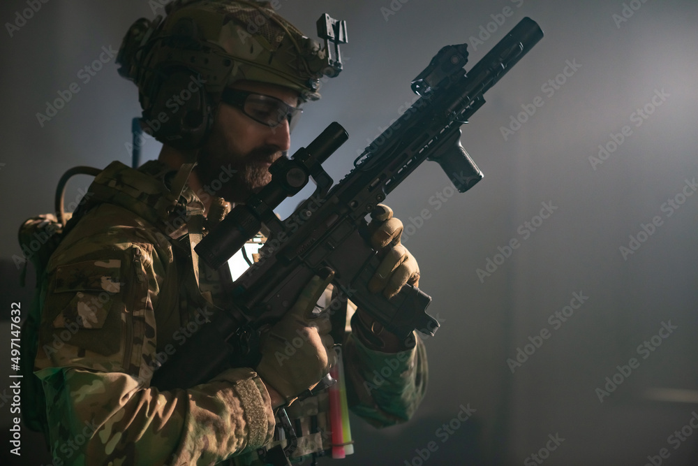 Attentive bearded man in military equipment in dark room. Man in uniform holding machine gun, getting ready for combat. Military, army, airsoft concept Attentive bearded man in military equipment in dark room. Man in uniform holding machine gun, getting ready for combat. Military, army, airsoft concept