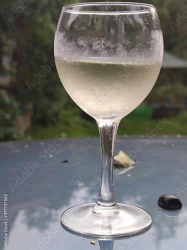 Cold white wine in a glass. In a transparent, misted glass goblet with a stem, cold white wine is slightly yellowish in color. The glass is on the green table. Wine with small air bubbles.