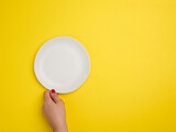 female hand holding a white empty round plate on a yellow background, top view