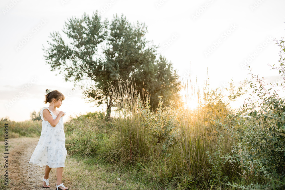 Tender little girl wearing natural white dress with wildflower motiv with wild carrot flowers in hair walking on the path in the field at summer, outdoor lifestyle backlit photo.