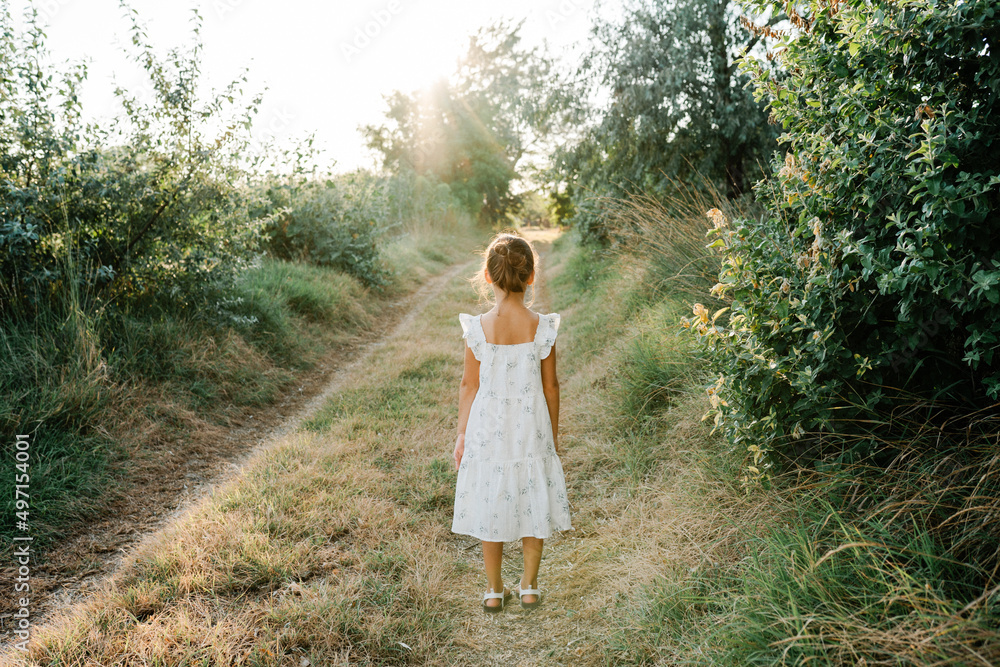 Tender little girl wearing natural white dress with wildflower motiv standing on the path in the field at summer, outdoor lifestyle backlit photo.