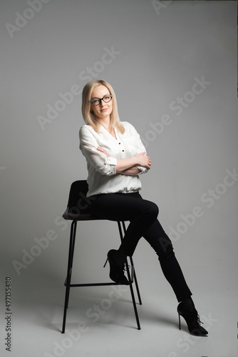 Beautiful young woman with glasses sitting on a chair in a studio with grey background. Sales agent, manager, agent, assistant, teacher, student