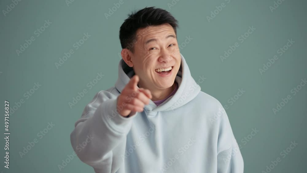 Smiling Asian man wearing hoodie pointing at the camera in the blue studio Smiling Asian man wearing hoodie pointing at the camera in the blue studio