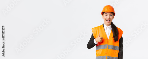 Supportive and pleased happy asian female chief engineer in safety helmet showing thumbs-up in approval, satisfied with construction work. Industrial woman in reflecive jacket inspect area photo