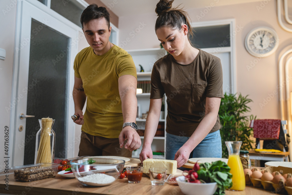 Two people young adult couple man and woman husband and wife or boyfriend and girlfriend preparing pizza food at home helping each other cutting and add ingredients and spices real people copy space Two people young adult couple man and woman husband and wife or boyfriend and girlfriend preparing pizza food at home helping each other cutting and add ingredients and spices real people copy space