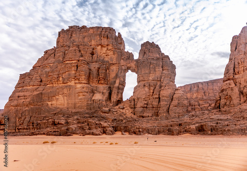 Tamesguida cathedral in Tadrart Rouge. Far tiny tourists climbing the natural window, sand and dry herbs with cloudy blue sky. Naturally sculpted rocky mountains of Tassili N'Ajjer national park.