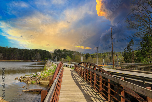 a metal and wooden rust colored bridge over a lake surrounded by lake water, green grass and lush green trees with blue sky and yellow clouds at Murphey Candler Park in Atlanta Georgia USA photo