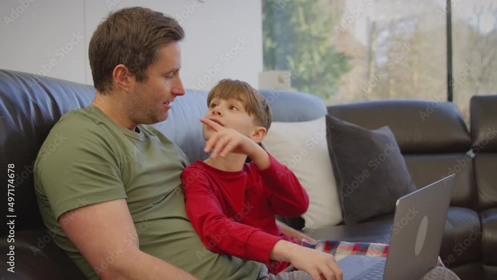 Father and son sitting on sofa at home in pyjamas together looking at laptop - shot in slow motion Father and son sitting on sofa at home in pyjamas together looking at laptop - shot in slow motion