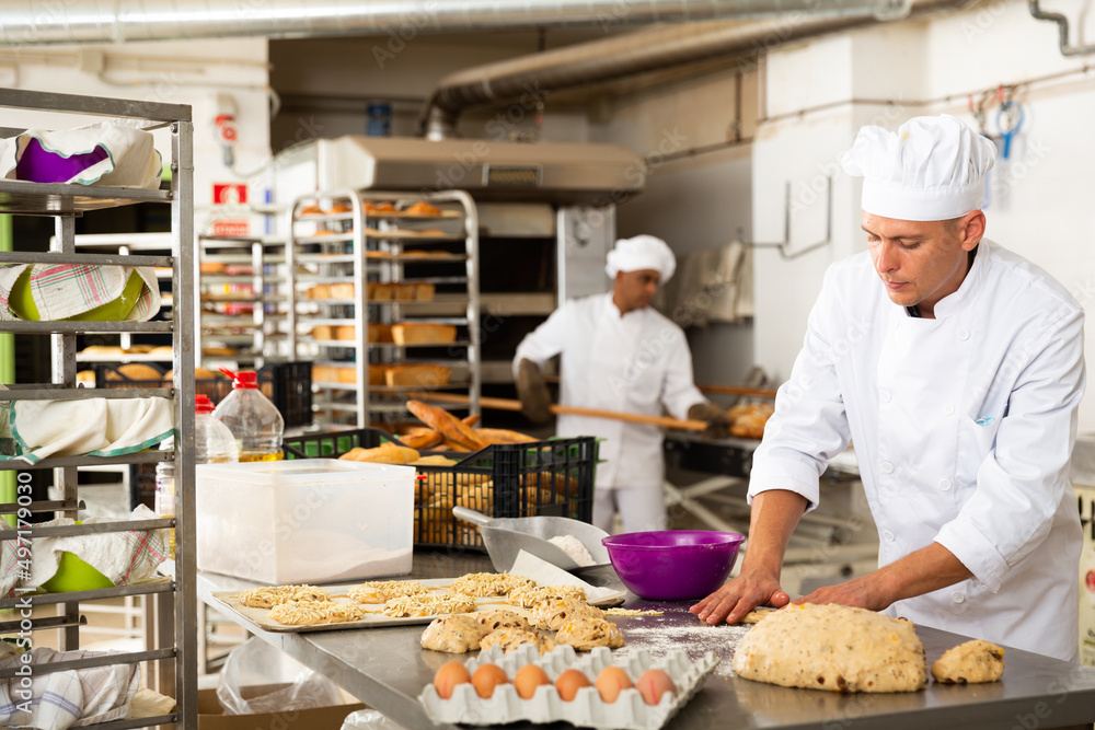 Working at bakery, male baker kneading dough and shaping baguettes on steel countertop in industrial kitchen