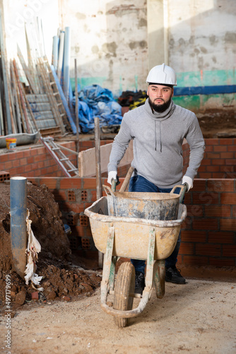 Young bearded man working at building site, carrying navvy wheelbarrow with bucket of construction mortar