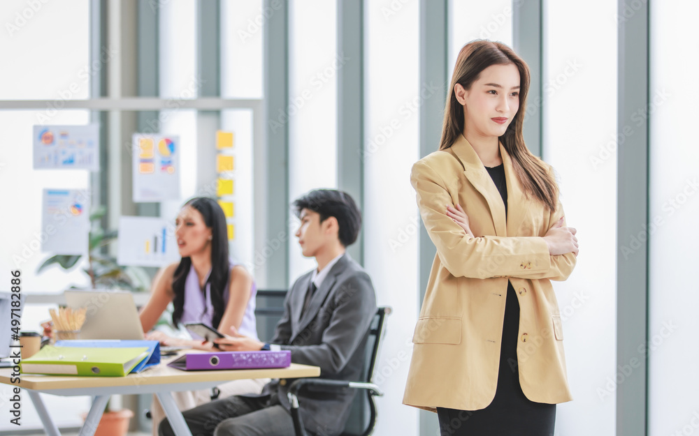 Asian young pretty happy businesswoman entrepreneur in formal business suit stand crossed arms smiling while male female businessman colleagues discussing brainstorming behind in office meeting room