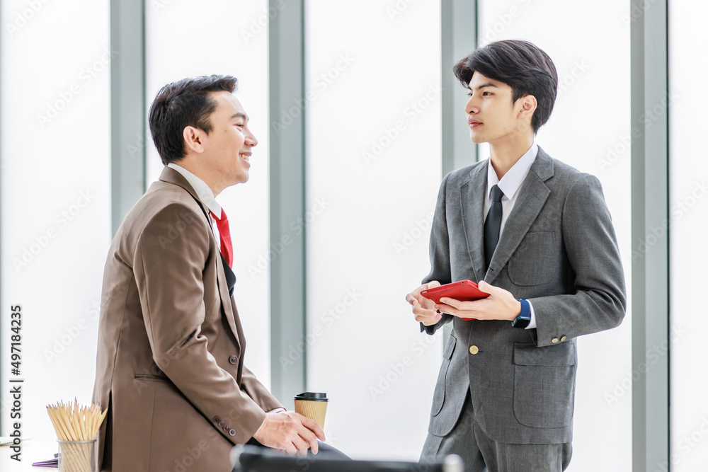 Asian male professional businessmen colleague in formal business suit take coffee break standing holding disposable paper cup and tablet computer talking have conversation together in front monitor Asian male professional businessmen colleague in formal business suit take coffee break standing holding disposable paper cup and tablet computer talking have conversation together in front monitor