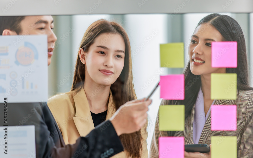 Asian happy male and female professional successful businessman and businesswoman colleagues partnership teamwork in formal business suit standing smiling look at post it note on glass board together Asian happy male and female professional successful businessman and businesswoman colleagues partnership teamwork in formal business suit standing smiling look at post it note on glass board together