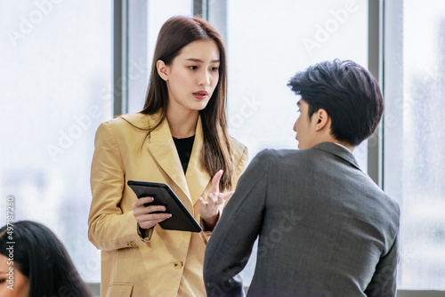 Asian young female businesswoman employee in formal business suit standing holding showing touchscreen tablet computer screen asking male businessman colleague for help and support in company office