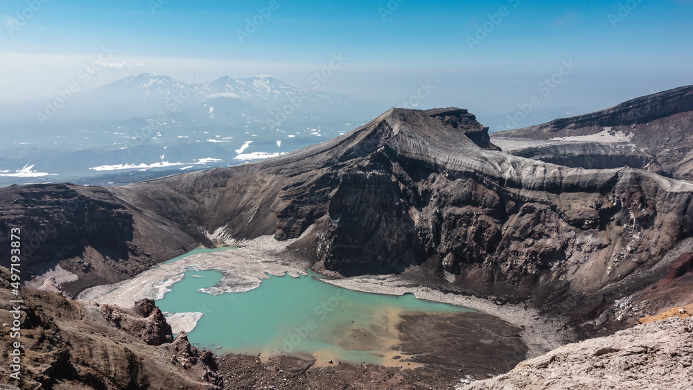 Amazing turquoise acid lake on top of an active volcano. Melted snow at the water's edge. The layered structure of the steep slopes of the crater is visible. Blue sky. Kamchatka. Gorely Volcano