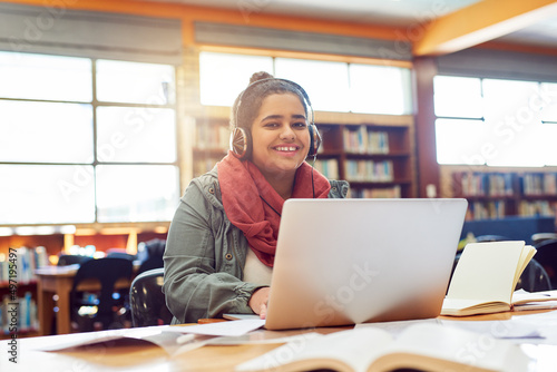 Music makes her perform. Portrait of a cheerful young female student working on a laptop while listening to music with her headphones inside of a library.