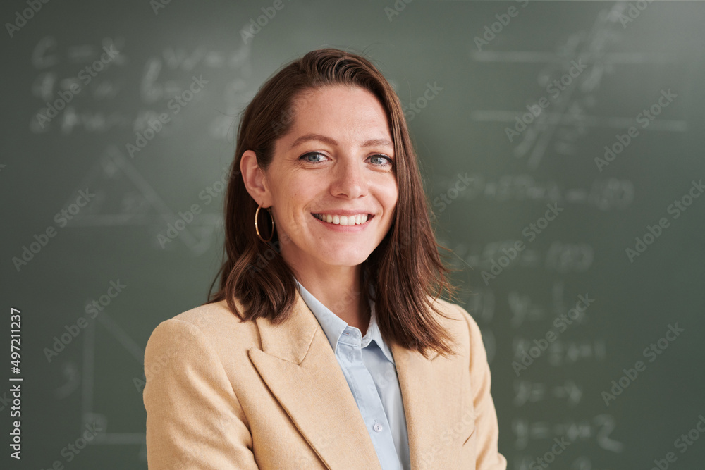 Portrait of young female teacher looking at camera and smiling while standing by blackboard in school classroom, copy space Portrait of young female teacher looking at camera and smiling while standing by blackboard in school classroom, copy space