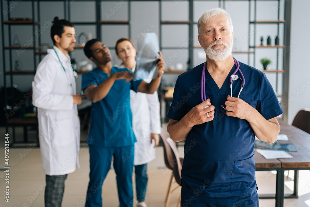 Obraz premium Portrait of confident senior aged surgeon in blue uniform standing in medical meeting office, looking at camera. Multiethnic team of physician working in background, discussing MRI scan of patient.