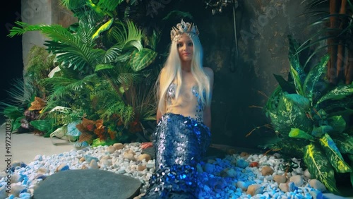 fantasy woman queen mermaid sits on floor in studio. Rain shower, green plant modern bathroom interior. Silver fish tail creative design costume fin, blonde nymph goddess girl princess, crown on head photo