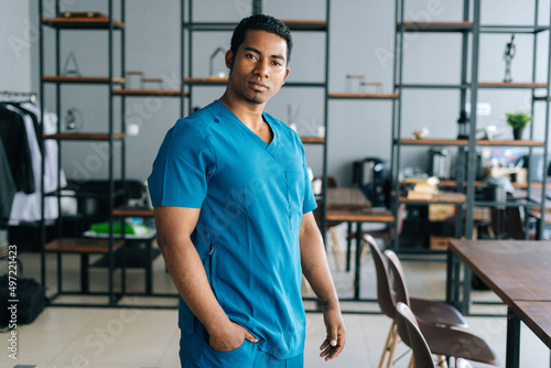 Portrait of confident African American man doctor wearing blue surgeon medical lab uniform standing in hospital office, looking at camera. Front view of experienced nurse male posing at workplace.