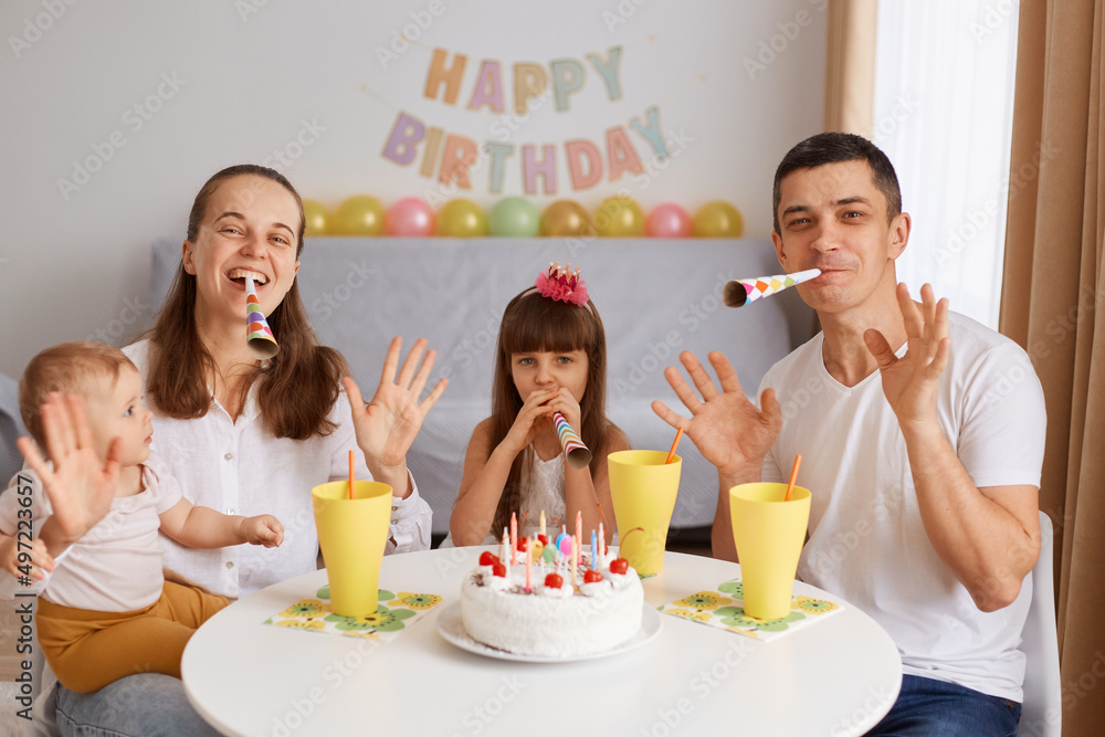 Portrait of delighted happy family sitting at the table and celebrating birthday, looking at camera with positive expressions, waving hands and blowing party cone. Portrait of delighted happy family sitting at the table and celebrating birthday, looking at camera with positive expressions, waving hands and blowing party cone.