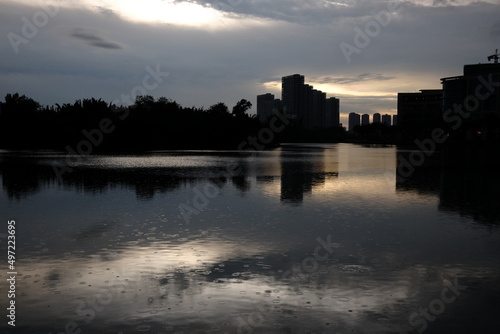 Surface of water in drops of rain, on the background is skyline and silhouette of the buildings and trees in sunset time. Rainy season theme. The cloudy sky is reflected in the river. 