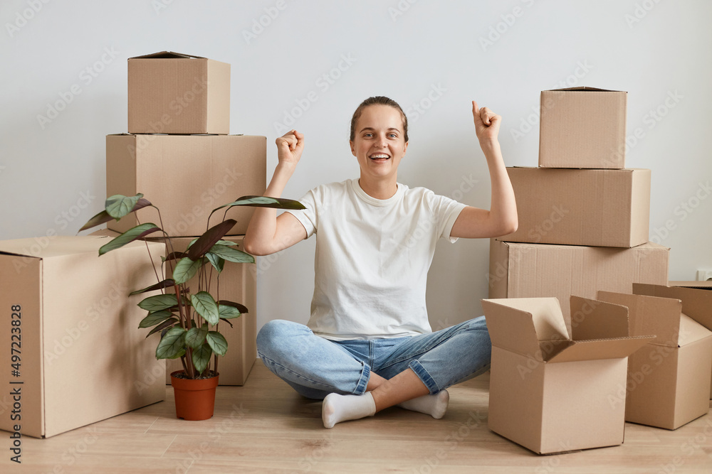 Extremely happy woman wearing white t shirt and jeans sitting on floor with crossed legs, celebrated her relocating to a new flat, clenched fists and looking at camera with toothy smile. Extremely happy woman wearing white t shirt and jeans sitting on floor with crossed legs, celebrated her relocating to a new flat, clenched fists and looking at camera with toothy smile.